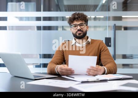 Jeune homme barbu avec des lunettes assis à un bureau dans un bureau moderne, tenant un document vierge, un ordinateur portable et des papiers sur la table, regardant directement la caméra avec une expression sérieuse Banque D'Images
