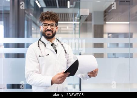 Jeune médecin masculin portant une blouse de laboratoire et un stéthoscope, tenant des documents médicaux tout en se tenant debout dans une clinique ou un hôpital moderne, regardant la caméra et souriant Banque D'Images