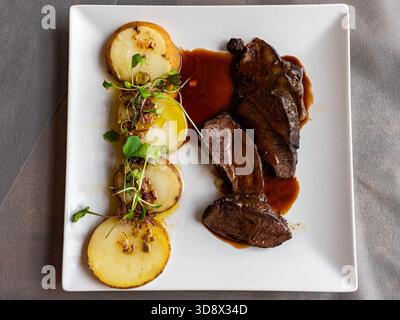 Vue de haut en bas d'un plat gastronomique : viande tendre tranchée servie avec une sauce brune riche, pommes de terre décorées et micro-légumes verts sur une assiette carrée. Banque D'Images