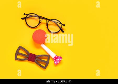Visage drôle fait de nez de clown, lunettes, noeud papillon et sifflet de fête sur fond jaune Banque D'Images