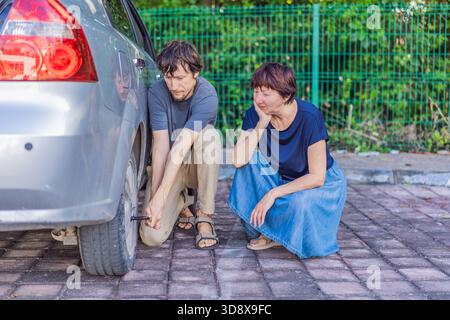 Jeune homme aidant une femme plus âgée en changeant la roue de la voiture alors qu'elle est assise à proximité, montrant soutien, gentillesse et assistance routière réelle. Famille Banque D'Images