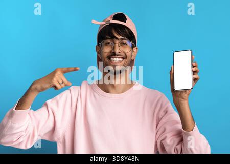 Jeune homme portant Pink Sweater et Cap montre joyeusement smartphone sur fond bleu dans un moment joyeux d'expression Banque D'Images