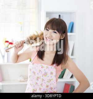 Heureuse femme au foyer asiatique avec tablier, main tenant un plumeau et souriant. Jeune femme à l'intérieur vivant le mode de vie à la maison Banque D'Images