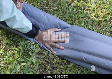 Personne concentrée avec la main sur un matelas pneumatique, préparant l'équipement pour une aventure de camping en plein air. Coussin de couchage gris dégonflant sur herbe verte pour hiki Banque D'Images