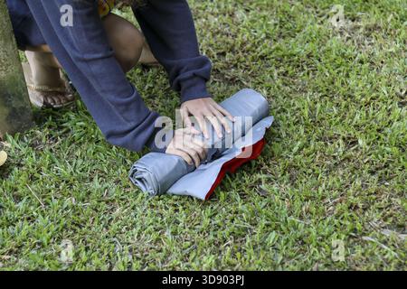 Personne concentrée emballant l'équipement de camping, roulant soigneusement un matelas de couchage gris ou un matelas pneumatique pour tente. Une activité de plein air sur herbe verte nécessitant du dilige Banque D'Images