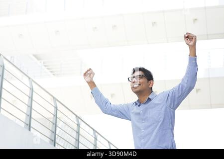 Homme d'affaires indien asiatique prospère avec les armes en place célébrant sa victoire, immeuble de bureaux moderne comme arrière-plan Banque D'Images
