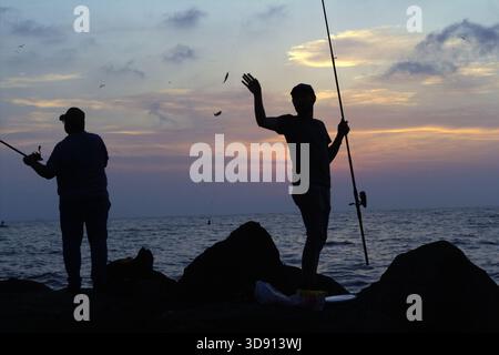 Pêcheur enthousiaste agitant sur Rocky Shore au coucher du soleil. Une silhouette d'un pêcheur se tient sur un rocher, agitant avec excitation tout en tenant sa pêche Banque D'Images