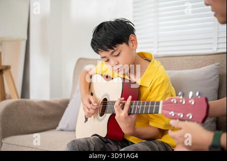 Gros plan de l'enfant élémentaire asiatique jouant une guitare classique sur le canapé dans leur salon de la maison ou école de musique. Instrument de musique, activité ludique. Banque D'Images