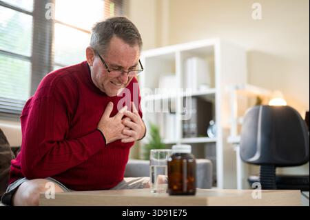 Vieil homme blanc caucasien portant des lunettes ayant une crise cardiaque ou un arrêt cardiaque assis sur un canapé dans un salon. Maladie et maladie. Problèmes de santé. Banque D'Images