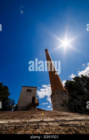 Une vue puissante et à faible angle d'une grande cheminée en brique et d'un bâtiment en béton délabré, encadré par un ciel bleu ensoleillé et vif avec un soleil proéminent Banque D'Images