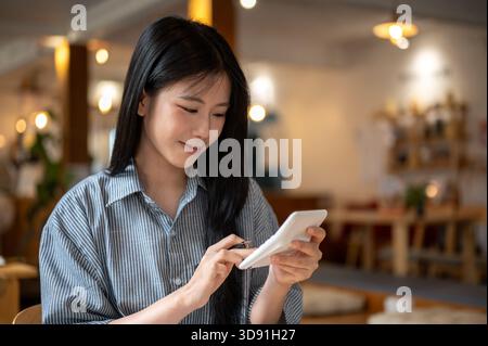 Femme asiatique souriante tenant et utilisant une calculatrice blanche tout en étant assis à la table en bois dans le café. Revenus et dépenses. Gestion de l'argent. Banque D'Images