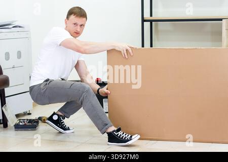 Homme accroupissant et poussant une lourde boîte en carton dans le bureau. Jeune homme fort en vêtements décontractés squattant et poussant une grande feuille de carton à travers le plancher indo Banque D'Images