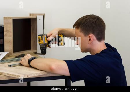 Profil de meubles de forage d'homme avec concentration. Vue latérale concentrée d'un jeune homme utilisant une perceuse électrique sur une partie de meuble en panneau de particules Banque D'Images
