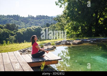 Femme mature méditant dans la position yoga lotus par étang de natation naturel dans le jardin de l'arrière-cour. Mode de vie sain grâce à la pleine conscience quotidienne et à la méditation Banque D'Images