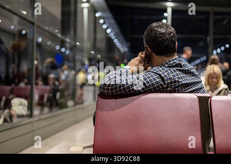 homme en chemise à carreaux est assis dans une zone d'attente d'aéroport ou de gare la nuit, regardant par une grande fenêtre une jeune femme souriante dehors. séparation, Banque D'Images
