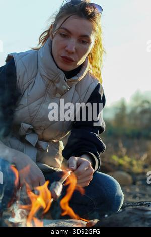 Femme aux cheveux blonds lâches et lunettes de soleil sur la tête toast une guimauve sur un bâton de bois au-dessus d'un petit feu de camp au milieu de l'herbe d'automne et des rochers (108) Banque D'Images