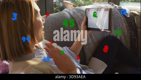Agitant femme chinoise tenant la tablette assise sur le canapé à la maison dans un pull beige avec des chiffres superposés Banque D'Images
