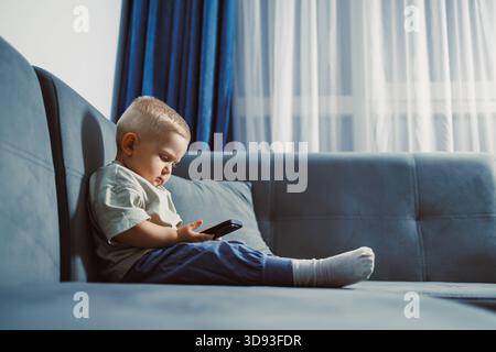 Un petit garçon est assis confortablement sur un canapé bleu, regardant attentivement son smartphone. La lumière chaude du soleil filtre à travers les rideaux, créant une atmosphère calme Banque D'Images