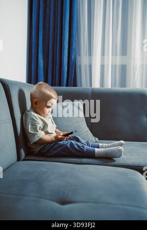 Un tout-petit s'assoit confortablement sur un canapé bleu doux, concentré sur un smartphone. La lumière du soleil coule à travers des rideaux transparents, remplissant l'espace de vie confortable. Banque D'Images