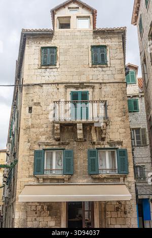 Une grande façade en pierre avec des volets turquoise, un petit balcon et un auvent beige le long d'une étroite rue européenne. Murs altérés et charme rustique évoquent Banque D'Images