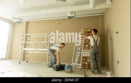 Deux ouvriers de la construction sont vus installer des échafaudages et organiser des outils dans un espace de bureau nouvellement construit. Les murs sont de couleur claire et le sol Banque D'Images
