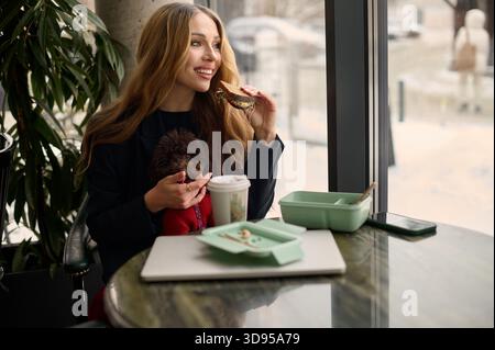 Jeune femme assise dans un café confortable, souriante tout en parlant au téléphone et en profitant d'un déjeuner à emporter. Banque D'Images