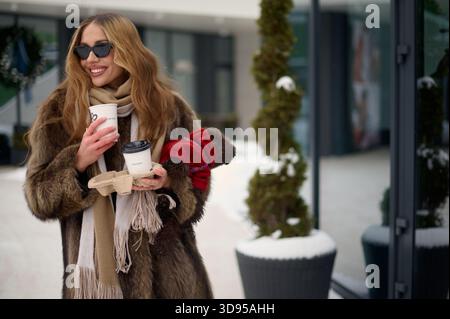 Heureuse femme à la mode dans des lunettes de soleil et un manteau de fourrure tenant deux tasses à café à emporter et un petit chien brun habillé dans un pull rouge tout en se tenant debout ou Banque D'Images