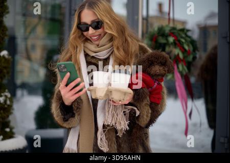 Heureuse femme à la mode dans des lunettes de soleil et un manteau de fourrure tenant deux tasses à café à emporter et un petit chien brun habillé dans un pull rouge tout en se tenant debout ou Banque D'Images