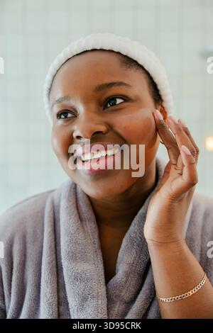 Femme afro-américaine lissant la crème dans la joue avec le bout des doigts dans la salle de bains portant un peignoir Banque D'Images
