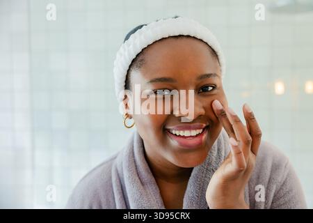Femme afro-américaine souriante, touchant la joue dans la salle de bain portant bandeau, peignoir, boucles d'oreilles Banque D'Images
