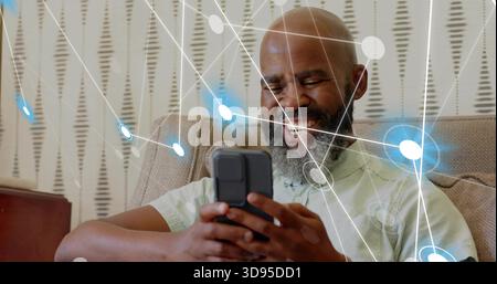 Homme souriant en chemise verte tenant un smartphone, naviguant sur un canapé beige à la maison, superpositions de réseau Banque D'Images