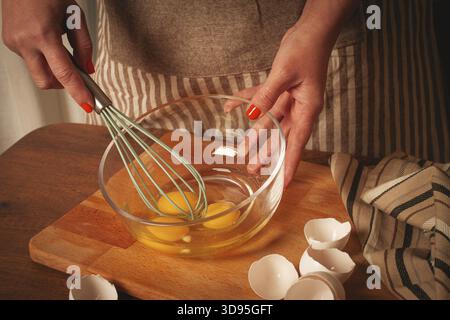 Les mains mélangent des œufs frais dans un bol en verre avec un fouet. Les coquilles d'œufs sont dispersées sur la surface en bois, indiquant la préparation d'un repas savoureux Banque D'Images