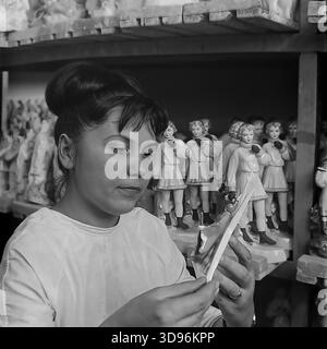 Slavyansk, Ukraine, URSS. 1968. Photo d'archives en noir et blanc montrant une employée du Slavyansk Ceramic combine effectuant le contrôle de la qualité des produits en porcelaine. La femme dans un manteau de travail blanc inspecte de près une petite figurine dans ses mains. Derrière elle, des étagères en bois sont garnies de rangées de statuettes identiques représentant des filles en manteaux et manteaux d'hiver. L'image illustre la production de masse de souvenirs de consommation et l'attention portée aux détails dans le Donbass pacifique avant la guerre. Banque D'Images