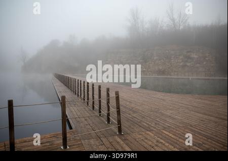 Une scène brumeuse avec un quai en bois s'étendant au-dessus d'une étendue d'eau calme, entouré de collines brumeuses et d'arbres. Zakrzowek, Cracovie Banque D'Images