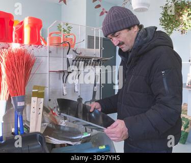 Homme asiatique mature en veste chaude et chapeau en tricot examine les pelles et les outils agricoles à l'intérieur du pavillon du marché ou du centre de jardinage, se préparant au travail ou Banque D'Images