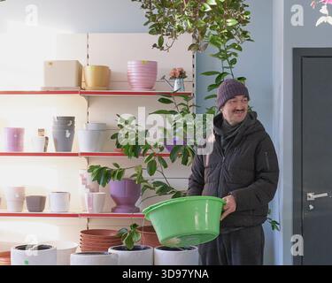 Homme asiatique mature souriant se tient dans le pavillon du marché, tenant un grand bassin en plastique vert tout en étant entouré de pots de fleurs colorés et de plantes d'intérieur sur sh Banque D'Images
