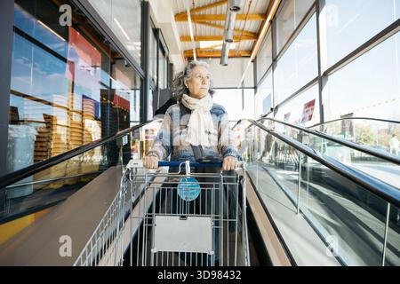 Femme senior déplaçant le chariot dans un escalier roulant dans un hall moderne de supermarché Banque D'Images