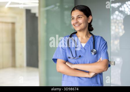 Professionnelle de la santé debout, les bras croisés, souriant tout en regardant loin, portant des gommages bleus et un stéthoscope dans un hôpital ou une clinique s. Banque D'Images