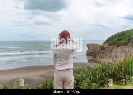 Femme asiatique en tissu décontracté avec chapeau appréciant sur la côte ouest par l'océan pacifique près de Muriwai Gannet Colony à Auckland, Nouvelle-Zélande Banque D'Images