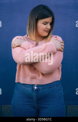 Jeune femme confiante en pull rose s'embrassant, debout contre un mur bleu vif, symbolisant l'amour de soi et la positivité du corps. Banque D'Images