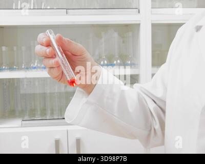 Technicien de laboratoire tenant un tube à essai rouge, gros plan sur la main gantée inspectant le liquide contre l'étagère de verrerie, Banque D'Images