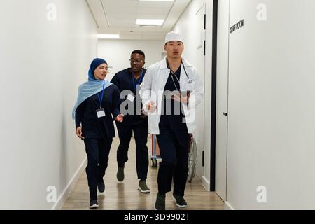 Personnel médical diversifié marchant dans le couloir de l'hôpital, médecin asiatique tenant un comprimé, portant un manteau blanc Banque D'Images