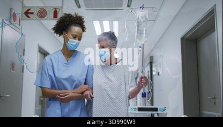 Infirmière ambulante dans des gommages bleus aidant le patient âgé portant une blouse et un masque d'hôpital, tenant la pompe IV Banque D'Images