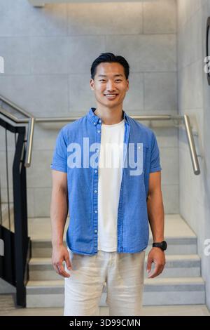 Homme asiatique debout sur des marches légères par des mains courantes en métal dans une cage d'escalier moderne portant une chemise bleu clair Banque D'Images