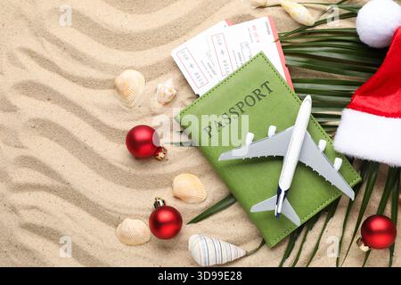 Vacances de Noël. Passeport avec billets, modèle d'avion, chapeau de Père Noël, coquillages, feuille de palmier et ornements sur la plage, pose à plat Banque D'Images