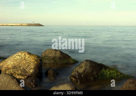 Gros plan longue exposition de Rocky Shoreline et de distant Breakwater gros plan en bas angle de roches sombres et moussues immergées dans l'eau claire et en mouvement doux Banque D'Images