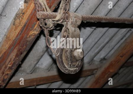 Une poulie rouillée est suspendue à des poutres en bois à l'intérieur d'une vieille structure avec un toit métallique. Le réglage montre des signes d'âge et d'usure. La poussière est visible en t Banque D'Images