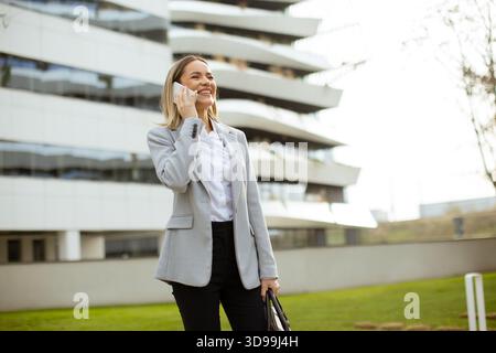 Une femme professionnelle sourit en parlant au téléphone devant un bâtiment contemporain. Elle porte un blazer et tient un sac, profitant d'un d ensoleillé Banque D'Images