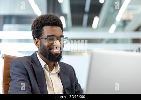 Homme d'affaires afro-américain souriant à l'ordinateur portable dans un bureau moderne et ensoleillé, axé sur le travail numérique confiant, gestionnaire professionnel dépeignant la productivité, le succès tech savvy Banque D'Images