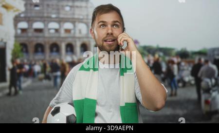 Homme tenant un ballon de football et un téléphone à l'oreille devant le bâtiment du colisée, portant une écharpe verte et une chemise blanche, regardant vers le haut ; anticipation de la fidélité à l'équipe Banque D'Images